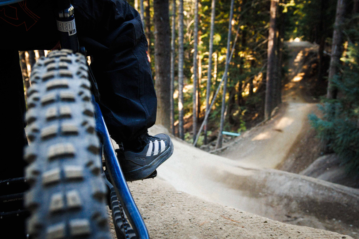 A close-up view of a mountain bike tire and the foot of a rider in specialized shoes, positioned on the bike's pedal. The background features a winding dirt trail through a forest, surrounded by tall trees and dappled sunlight.