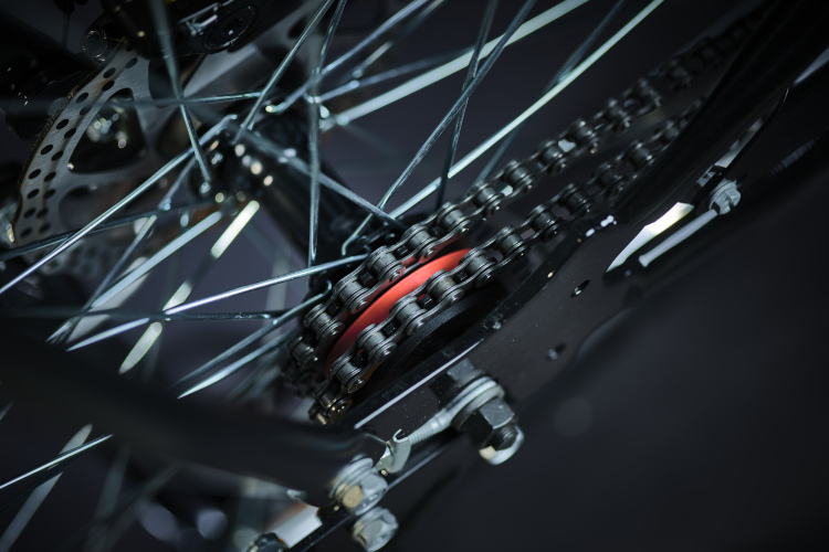 Close-up view of a bicycle's drivetrain, featuring the chain, sprockets, and rear wheel components. The image highlights the intricate details of the metallic chain links and gears against a dark background.