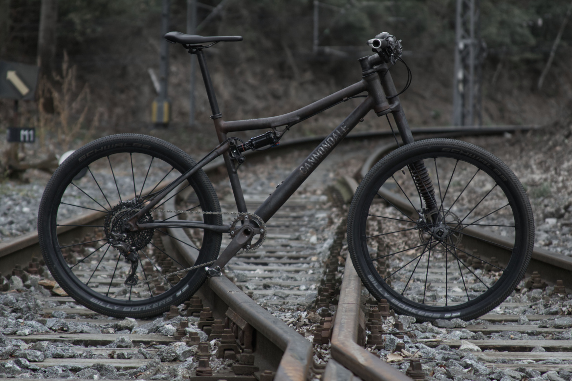 A mountain bike positioned on a set of railway tracks, featuring a dark frame and large, rugged tires. The setting is surrounded by scattered gravel and foliage, creating an outdoor atmosphere.