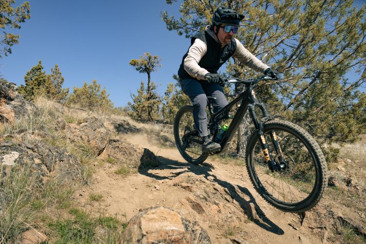 A mountain biker navigating a rocky trail during a sunny day, surrounded by trees and dust being kicked up from the ground. The rider is wearing a helmet and sunglasses, dressed in a sleeveless vest and athletic pants.
