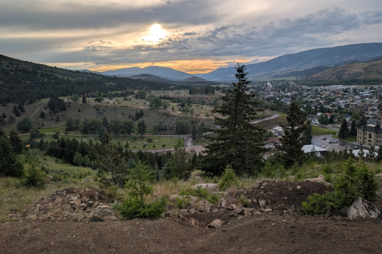 A panoramic view of a mountainous landscape at sunset, featuring rolling hills, a winding road, and a small town nestled in the valley. The sky is partly cloudy with hints of orange and yellow, casting a warm glow over the scene. Pine trees are visible in the foreground, adding to the natural beauty of the setting.