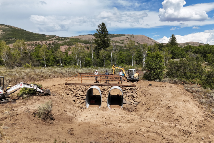 Construction site in a rural area showing two concrete archways built into a rocky structure, with workers standing on top. Excavators and construction machinery are on the ground, surrounded by sparse vegetation and mountains in the background under a partly cloudy sky.