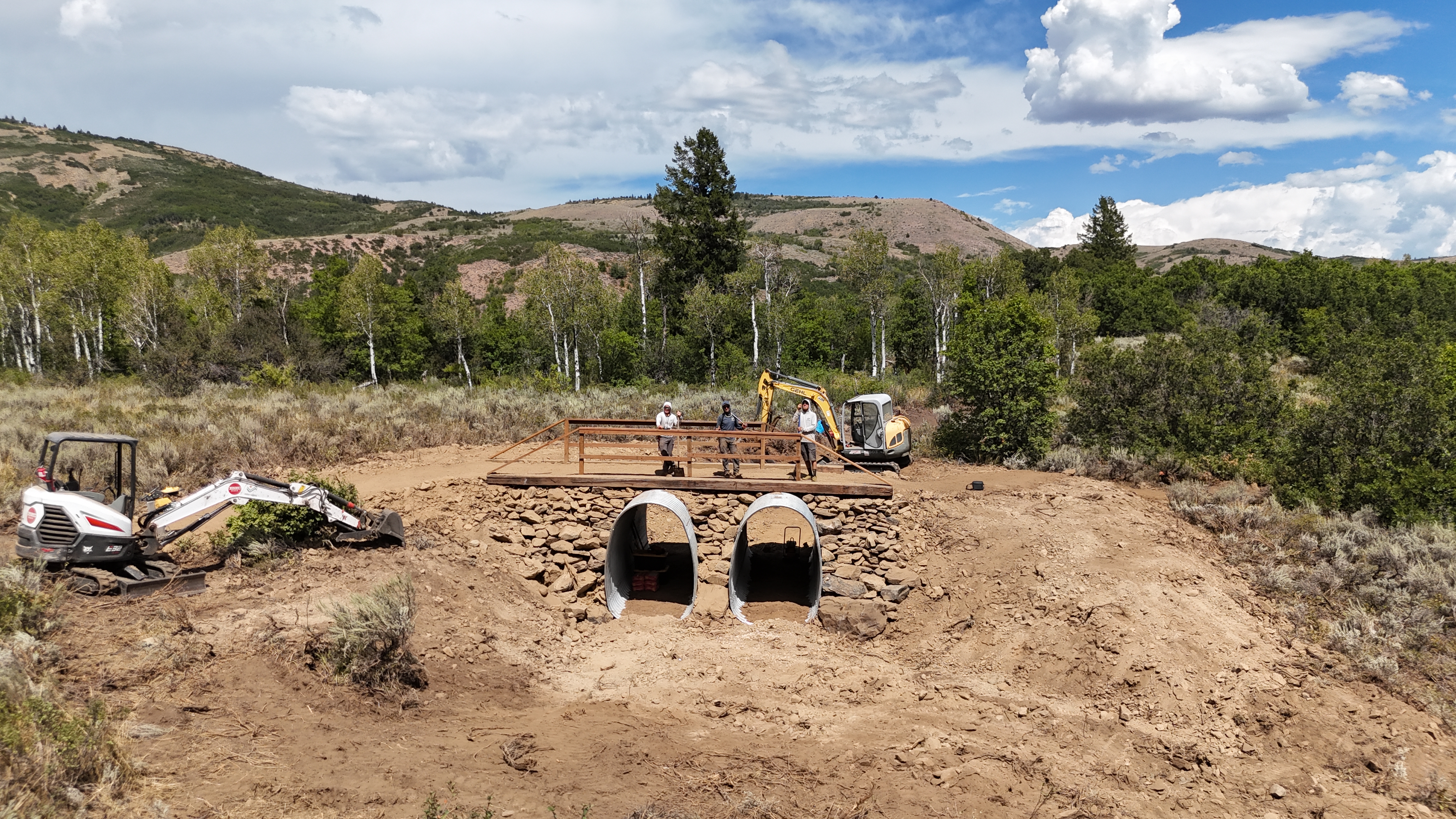 Construction site in a rural area showing two concrete archways built into a rocky structure, with workers standing on top. Excavators and construction machinery are on the ground, surrounded by sparse vegetation and mountains in the background under a partly cloudy sky.