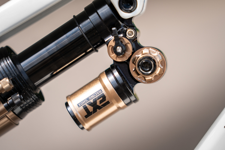 Close-up image of a bicycle shock absorber, featuring a combination of black and bronze components. The shock has detailed mechanical features, including dials and adjustment knobs, attached to a white bicycle frame. The background is softly blurred, emphasizing the shock absorber's intricate design.