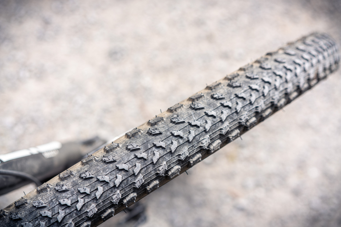 Close-up view of a bicycle tire with a textured tread pattern, resting on a gravel surface. The tire shows signs of wear, indicating use on rough terrain.