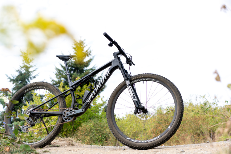 A black mountain bike with "Specialized" branding is positioned on a dirt path surrounded by greenery. The bike features thick tires and a sturdy frame, showcasing its design for rugged terrain. In the background, trees and foliage can be seen against a light sky.