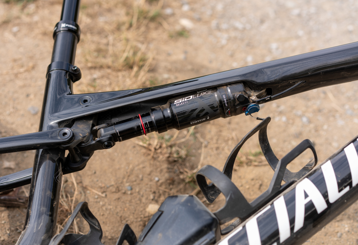 Close-up view of a mountain bike's suspension system, featuring a black shock with red and blue accents installed on a carbon frame, set against a dirt surface.