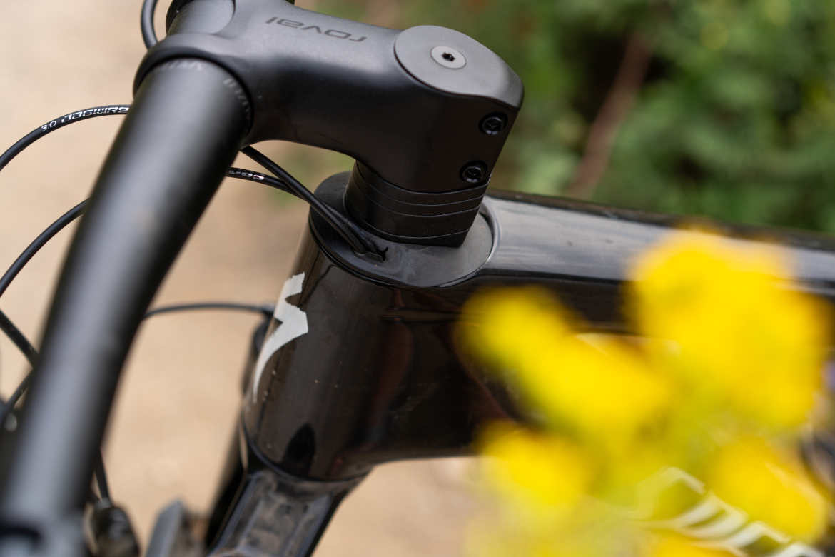 Close-up view of a bicycle's stem and handlebars, showcasing the black components and cables against a blurred background of yellow flowers and green foliage.