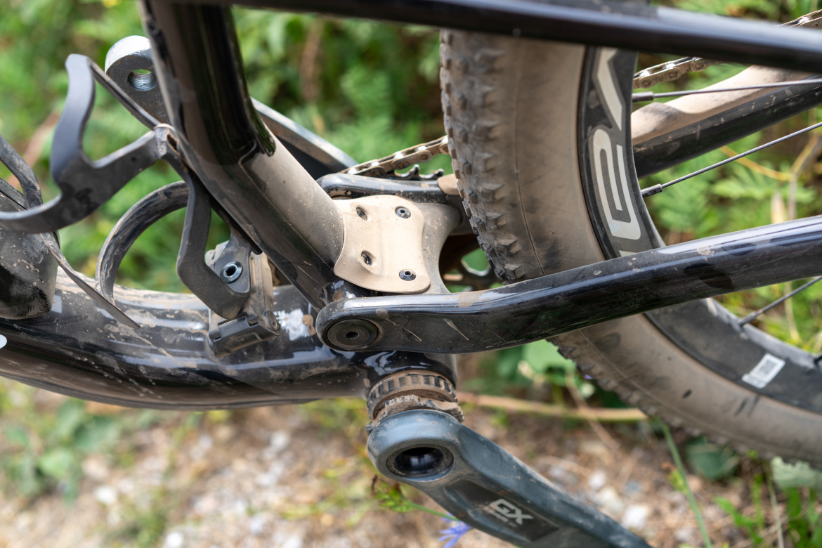 Close-up view of a mountain bike's drivetrain and bottom bracket area, featuring a black frame, crankset, and muddy tire, with greenery in the background.