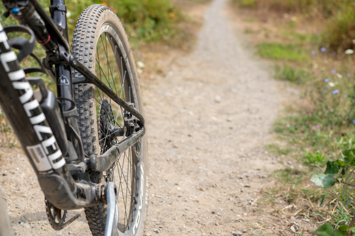 A close-up view of a mountain bike leaning on a gravel path, showcasing the rear wheel and part of the bike frame, surrounded by greenery and wildflowers in the background.