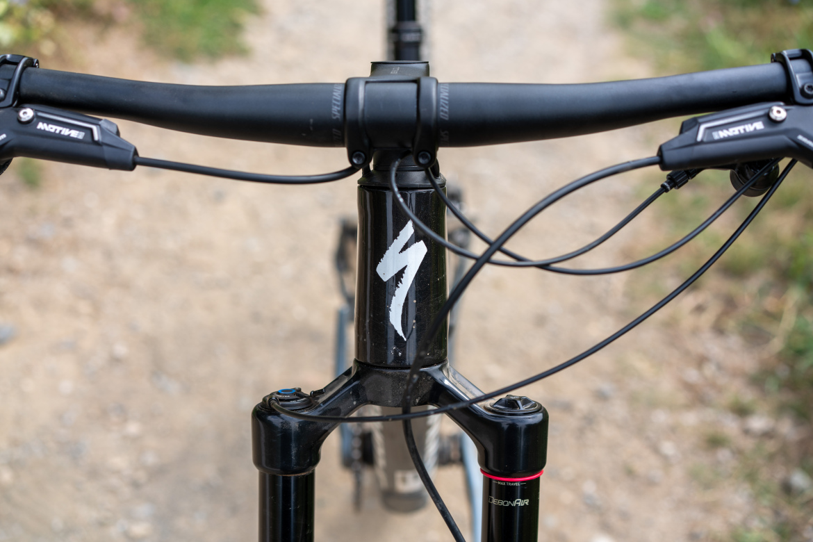 Close-up view of a bike's handlebars and stem, featuring a black frame with a white logo, black cables, and a fork with a red detailing. The background shows a gravel path surrounded by greenery.
