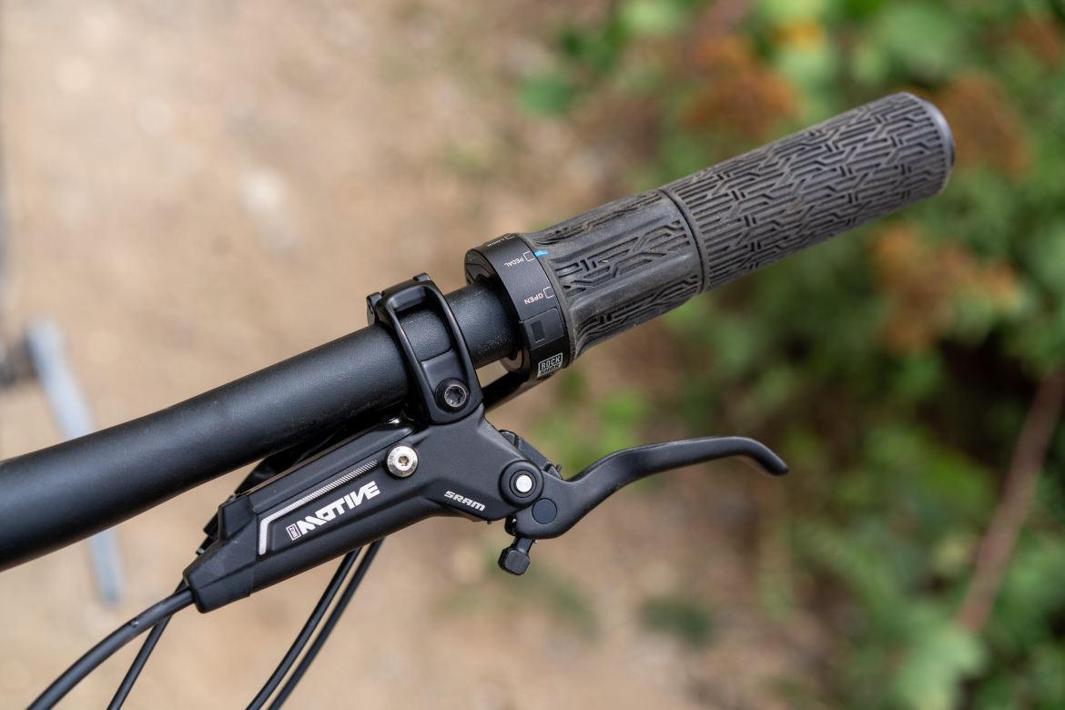 Close-up of a bicycle handlebar featuring a black grip and a SRAM brake lever. The handlebar is set against a blurred natural background, highlighting the bike's components.