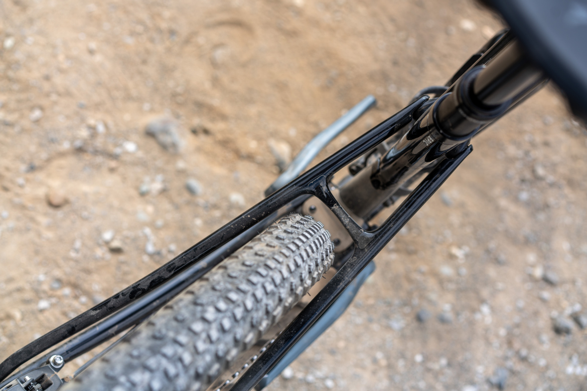 Close-up view of a mountain bike frame showing the rear tire and suspension system, with a gravel and dirt background.