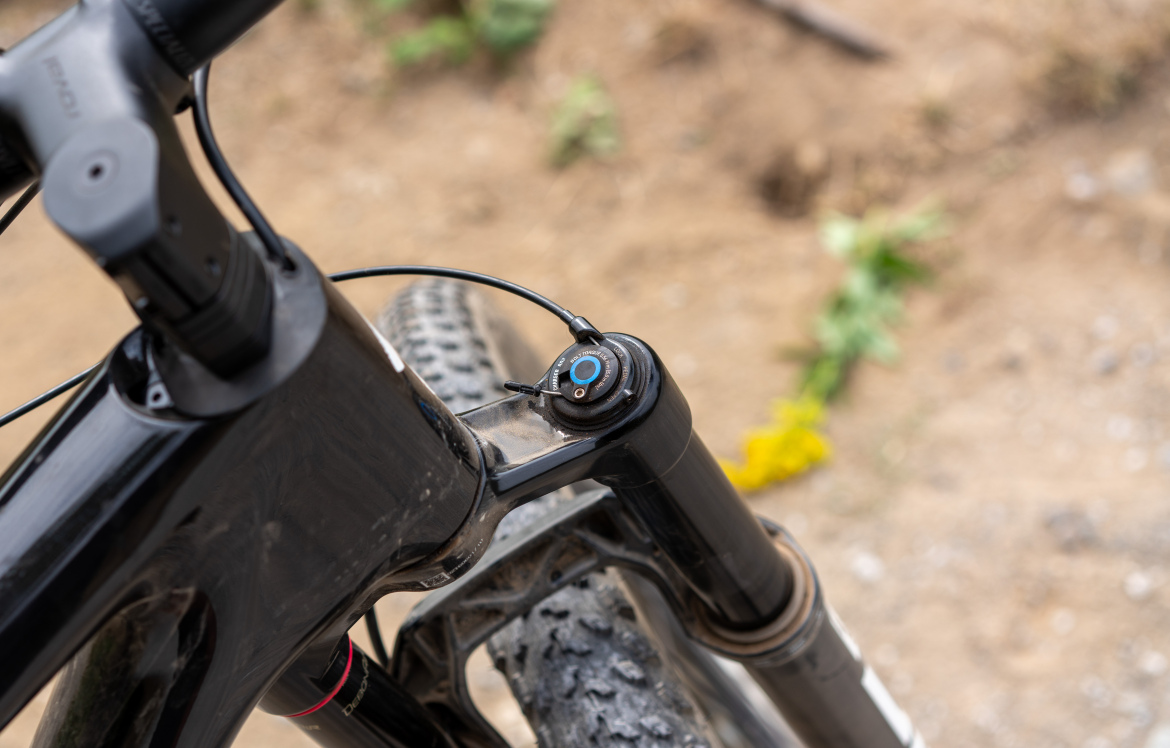 Close-up view of a mountain bike's head tube and fork, showcasing the black frame, cables, and a knob for adjusting the suspension settings. The background features dirt and greenery.