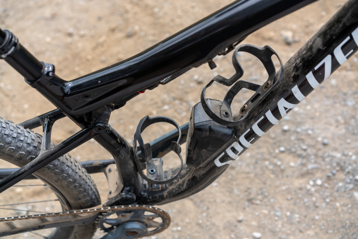 Close-up of a black mountain bike frame with visible water bottle cages, set against a gravel background. The bike is detailed with a logo and showing signs of use, including dirt and wear on the frame.