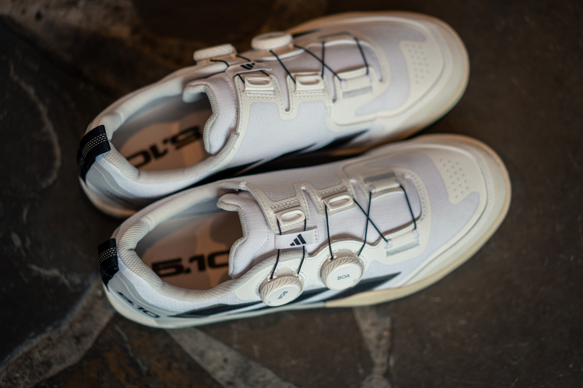 A pair of white athletic shoes with a sleek design featuring a Boa lacing system and a black accent. The shoes are positioned on a textured stone surface.