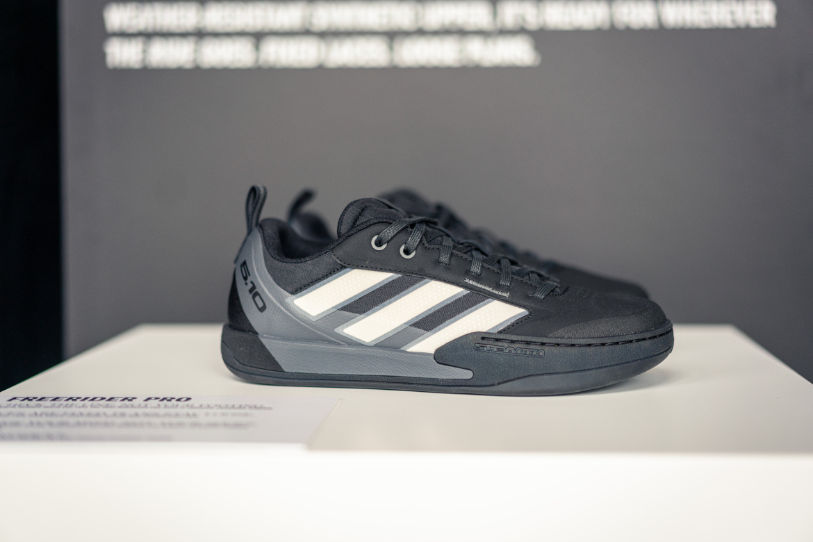 A pair of black and gray athletic shoes displayed on a white surface. The shoes feature a weather-resistant upper, a prominent logo, and three white stripes along the side. Background text is partially visible, discussing the shoe's features.