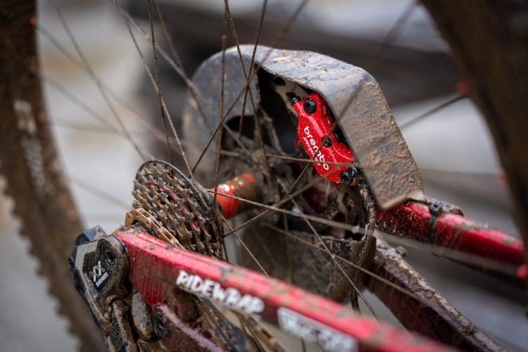 Close-up of a muddy bicycle drivetrain, featuring a red derailleur, gear cassette, and chain. The components are covered in dirt, highlighting the bike's use in off-road conditions. The visible branding on the frame reads "RIDEWRAP."