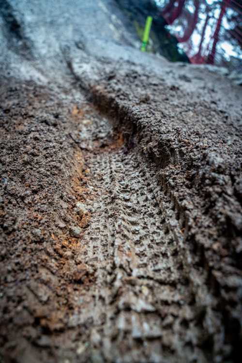 Close-up of a muddy surface showing tire tracks, with visible texture and variations in the earth. The background features hints of gravel and machinery parts.