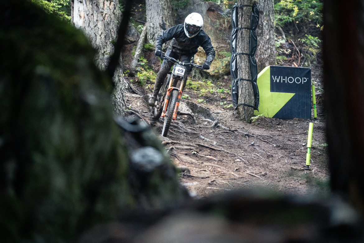 A mountain biker races along a dirt trail surrounded by trees, focused on navigating the terrain. The biker wears a helmet and protective gear, and the bike features an orange frame. In the background, a sign for "WHOOP" can be seen among the natural scenery. The trail shows signs of mud and roots, indicating a challenging course.