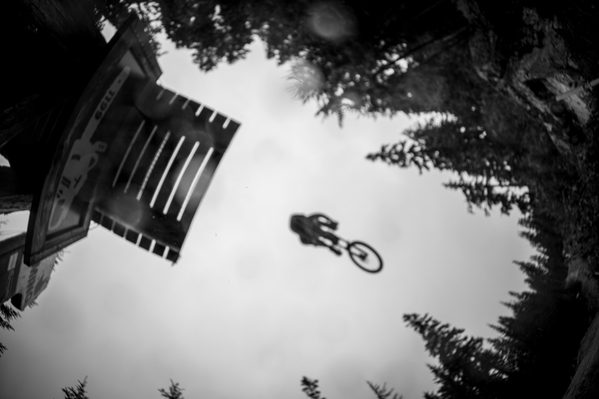 A black and white image capturing a mountain biker in mid-air as they jump off a ramp surrounded by pine trees. The ramp is partially visible in the upper part of the frame, indicating the height of the jump. The sky appears cloudy, adding a dramatic effect to the scene.