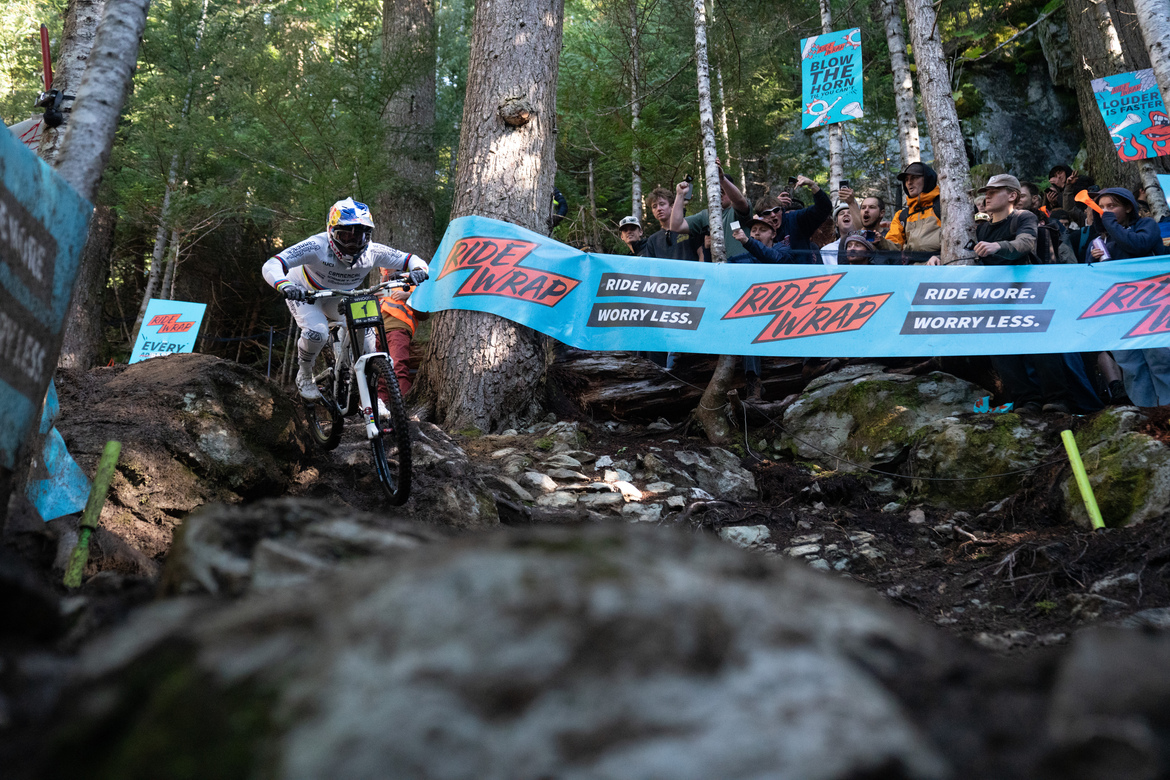 A mountain biker skillfully navigates a rocky descent in a forested area during a competition. Spectators cheer from the sidelines, surrounded by vibrant banners promoting a biking brand. The scene captures the excitement and energy of the event, with sunlight filtering through the trees.
