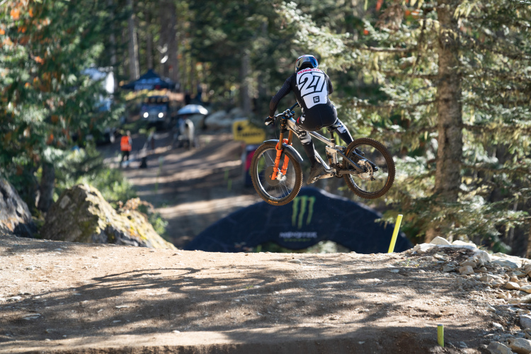 A mountain biker in a black and white outfit with the number 27 is performing a jump on a dirt track surrounded by trees. The background features a Monster Energy arch and spectators in the distance.
