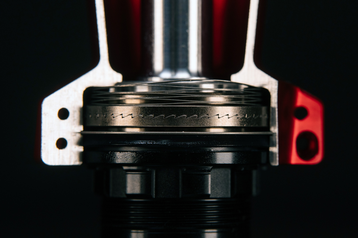Close-up view of a mechanical component, featuring a metallic structure with intricate details and textures, showing a combination of silver and black materials, likely part of a hydraulic or suspension system. The image highlights fine grooves and cutouts, set against a dark background, emphasizing the component's precision engineering.