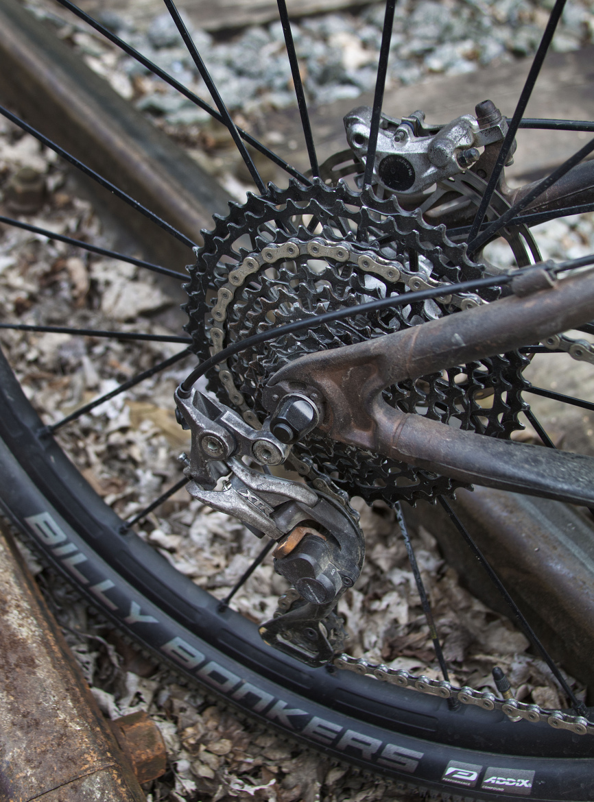 Close-up view of a bicycle's rear wheel cassette, derailleur, and chain, with leaves and gravel in the background. The focus is on the intricate mechanical components, highlighting the gears and chain arrangement.