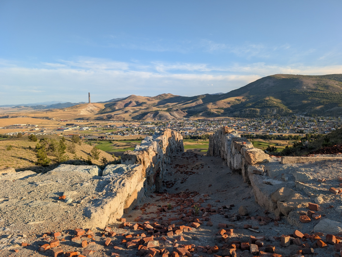 A panoramic view of a valley with a small town nestled below and hills in the background. In the foreground, a partially collapsed stone wall is visible, with scattered bricks and gravel. A tall smokestack rises from the landscape, and the sky is blue with a few clouds, indicating clear weather.