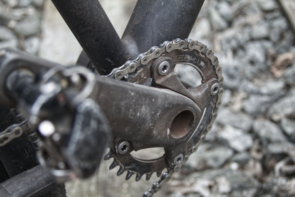 Close-up view of a bicycle's crankset and chain, showcasing the pedal attachment and the chain ring against a blurred, textured background of gray stones. The crank appears worn with dirt and age, highlighting its use in cycling.