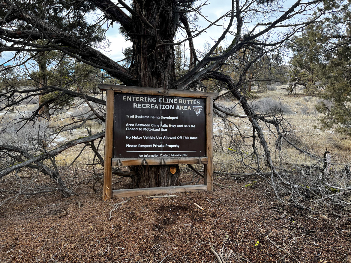 A wooden sign reading "Entering Cline Buttes Recreation Area" is positioned in front of a backdrop of trees and open land. The sign provides information about trail systems being developed, notes that the area between Cline Falls Highway and Barr Road is closed to motorized use, and requests that visitors respect private property.