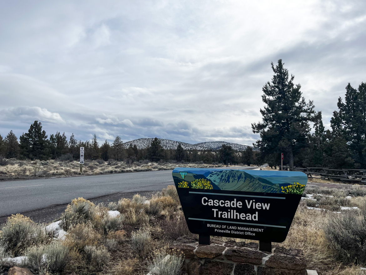 Sign marking the Cascade View Trailhead, featuring a colorful illustration of mountains and greenery. The surrounding area includes open land with sparse vegetation and trees, under a cloudy sky. A paved road is visible in the foreground, along with a parking area nearby.