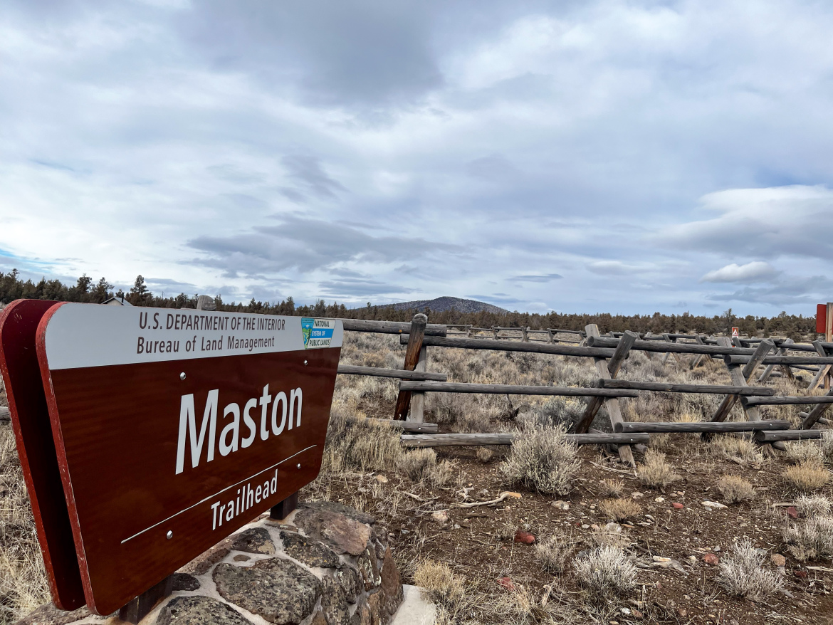 Sign for the Maston Trailhead, situated in a natural landscape with shrubs and a wooden fence, under a cloudy sky. The sign includes the U.S. Department of the Interior and Bureau of Land Management logos.