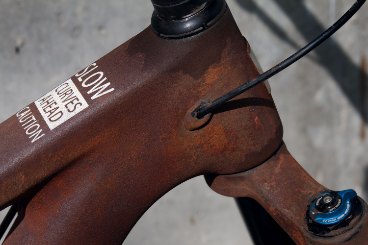 A close-up view of a rusted bicycle frame featuring a warning label that reads "SLOW CURVES AHEAD CAUTION." The frame shows signs of wear and corrosion, with a black cable running alongside the head tube and a blue adjustment knob visible on the side. The background is a blurred, textured surface.