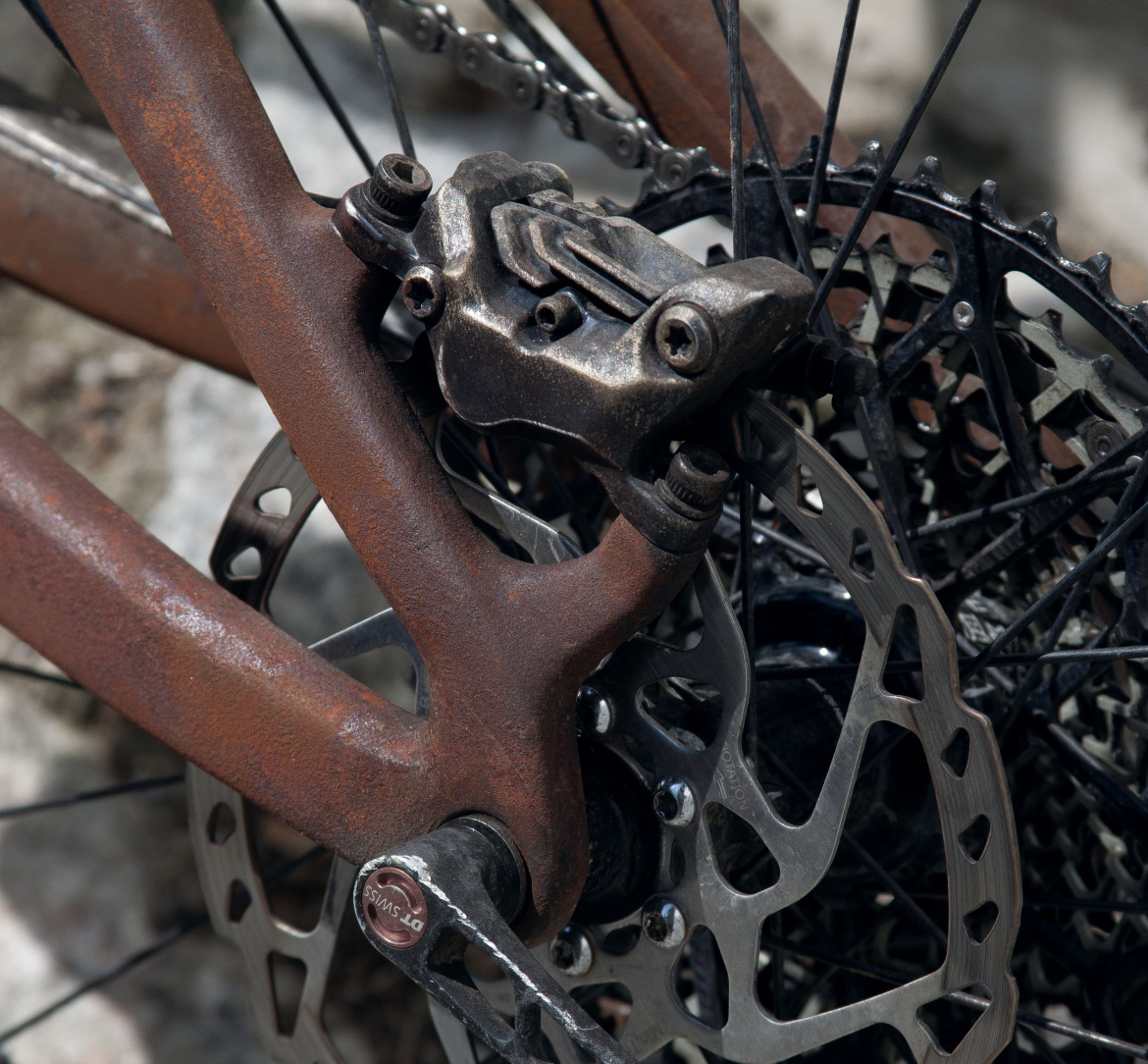 Close-up of a bicycle's rear braking system, featuring a rusted frame and a metallic brake caliper mounted near a disc brake. The intricate details of the components, including the chain and gearing, are visible in the background.