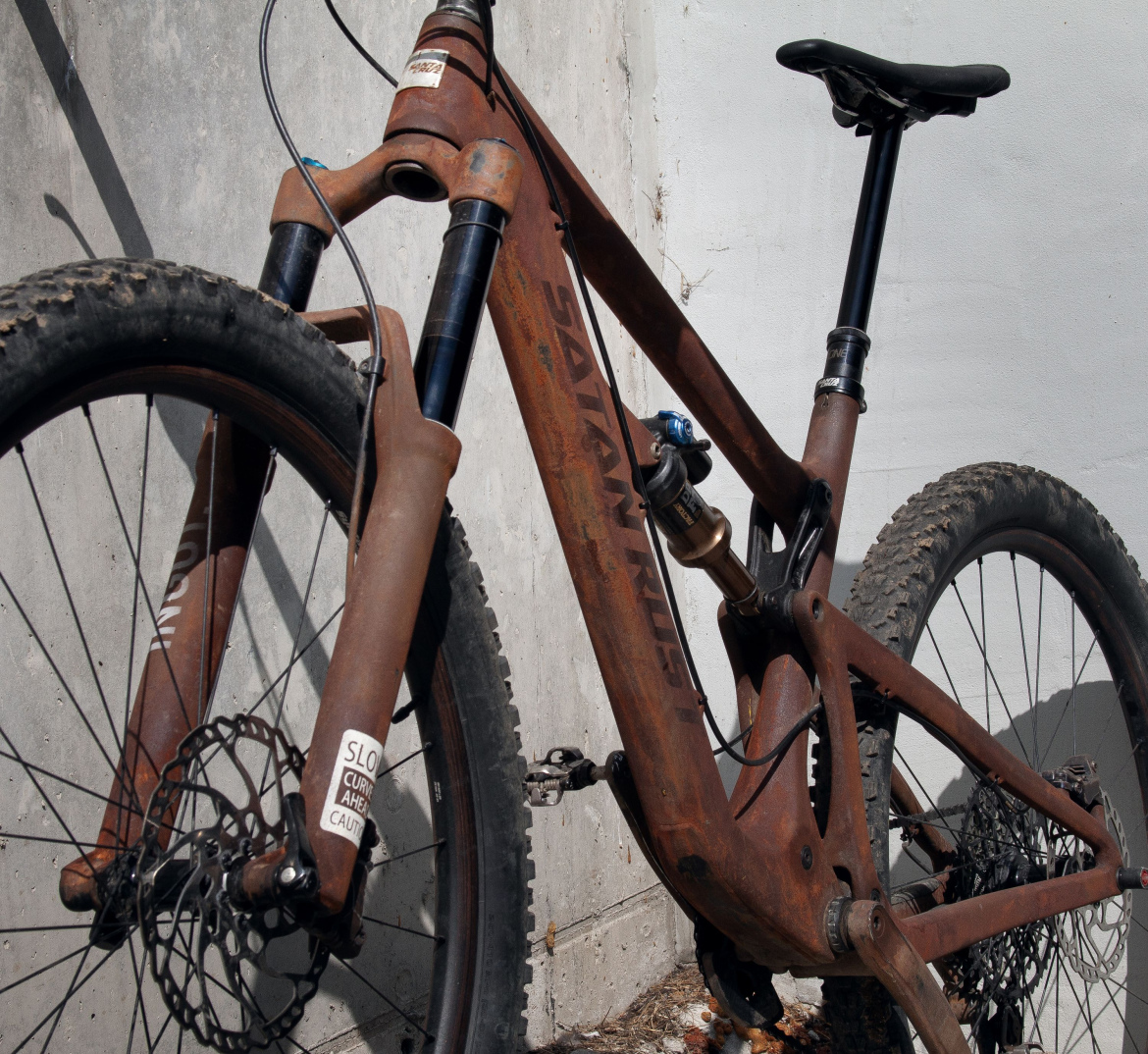 A close-up view of a rugged mountain bike with a weathered, reddish-brown frame, featuring a front suspension fork and disc brakes. The bike is leaning against a concrete wall, with dirt on the tires indicating outdoor use.