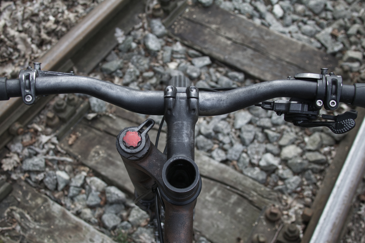 A top-down view of a bicycle handlebar with a dark metal frame, positioned above a railway track with scattered rocks and wooden planks in the background. The handlebar features a small red adjustment knob and a gear shifter on the right side.