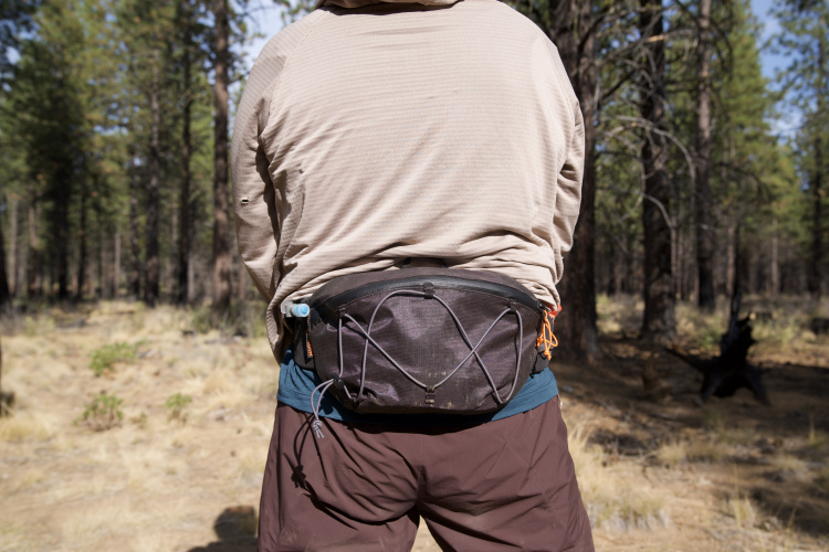 A person standing in a forest, wearing a light-colored long-sleeve shirt and brown shorts. They have a black waist pack secured around their waist, with some gear and a water bottle attached. The background features tall trees and grassy ground, indicating an outdoor setting.