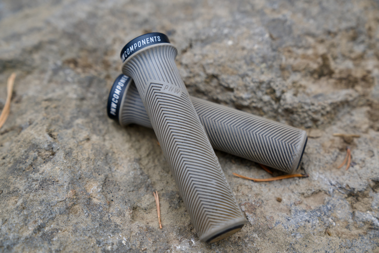 Two gray bicycle grips with a herringbone texture are resting on a rocky surface. The grips have black end caps with the text "PNW COMPONENTS" visible.