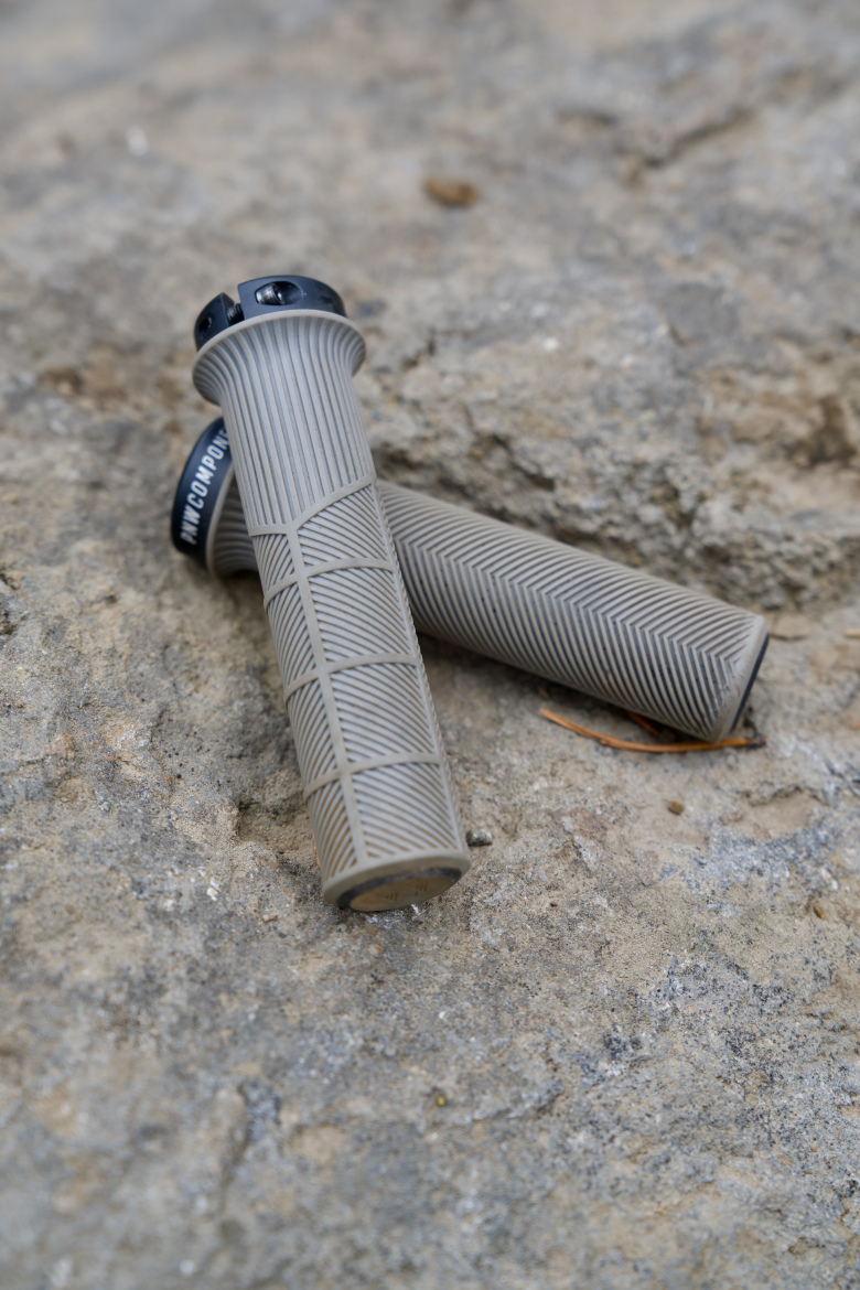 Two gray bicycle grips with textured surfaces are resting on a rocky surface. The grips have black end caps and feature a herringbone pattern for enhanced grip.