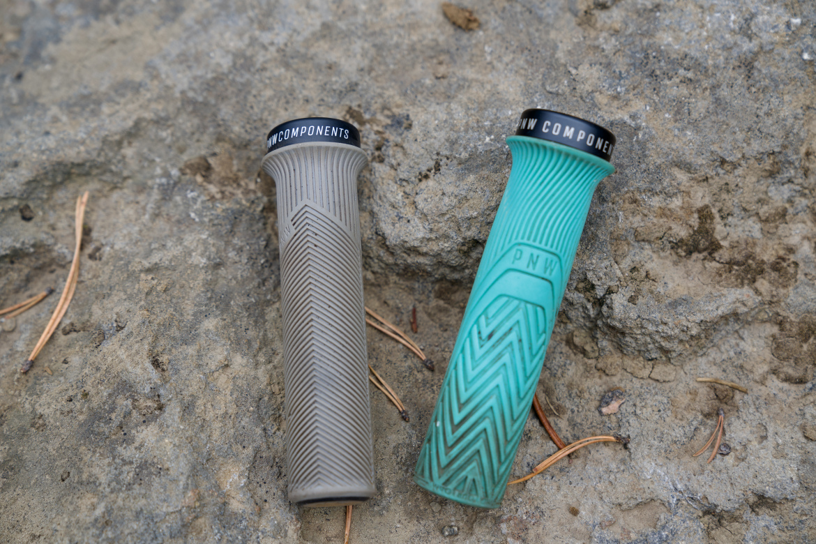 Two bicycle grips are displayed on a rocky surface. One grip is a muted gray color with a textured zigzag pattern, while the other is a bright teal with a similar design. Both grips feature a logo at the top. Pine needles are visible on the ground surrounding the grips.