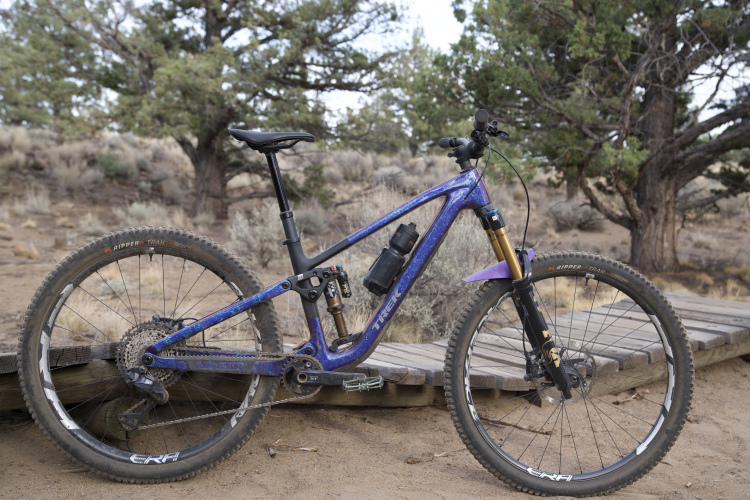 A purple mountain bike with a lightweight frame, front suspension, and drops of muddy dirt, parked on a wooden boardwalk in a rugged outdoor setting, surrounded by brush and trees.