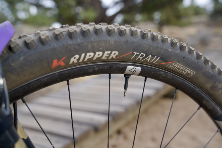 Close-up of a mountain bike tire with the text "K Ripper TRAIL" visible, showing its knobby tread and tubeless-ready design. The rim and a portion of the wheel's spokes are also visible, with a natural outdoor setting in the background.