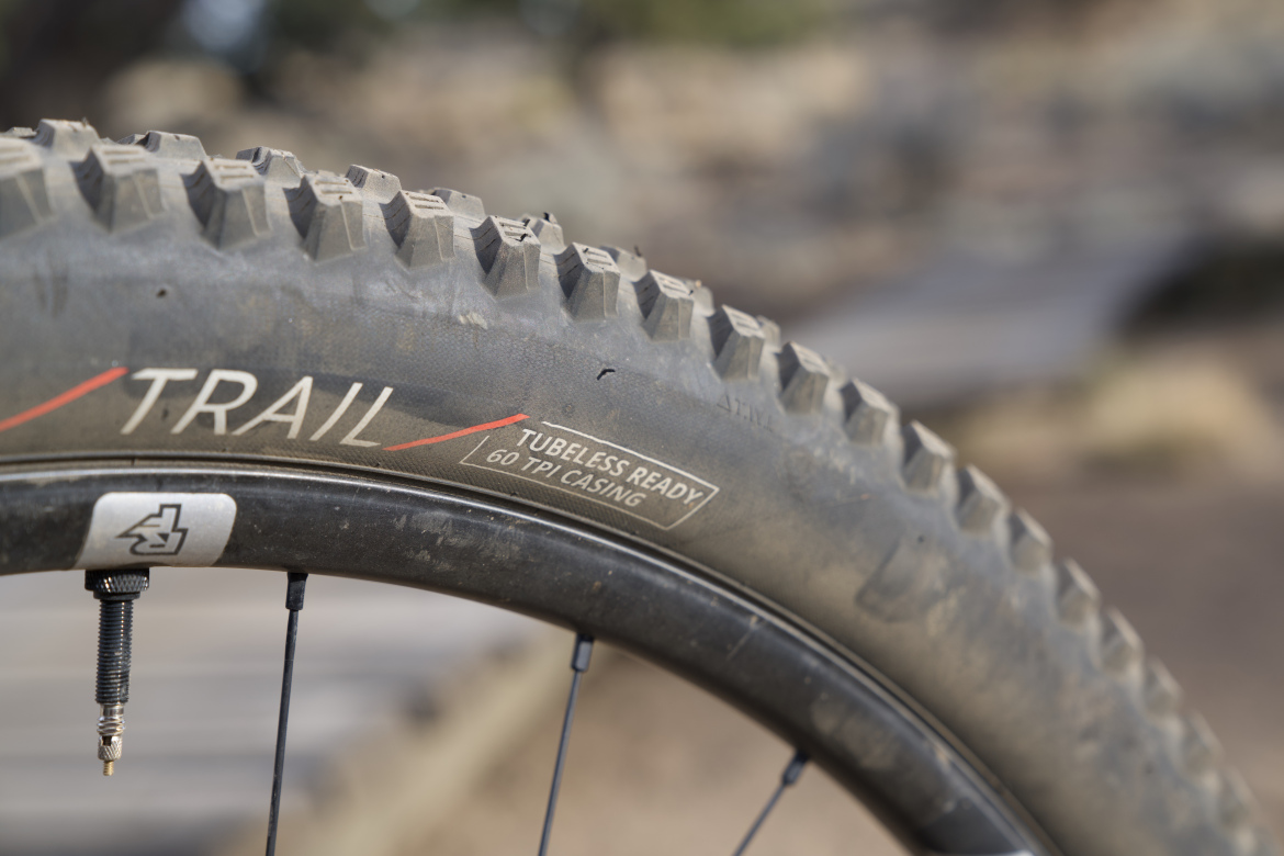 Close-up of a mountain bike tire labeled "TRAIL" with markings indicating it is tubeless ready and features a 60 TPI casing, showcasing the tread pattern and valve stem against a blurred outdoor background.