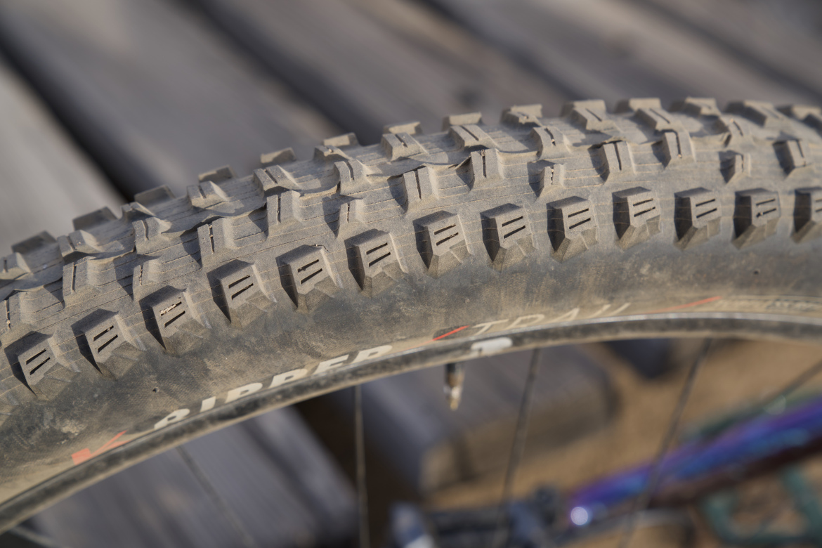 Close-up of a mountain bike tire showing its rugged tread pattern, designed for off-road traction, with dirt and dust visible on the surface. The background features wooden planks.