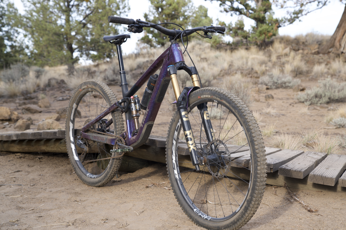 A close-up view of a mountain bike on a wooden bridge in a natural landscape. The bike features a purple and black frame, suspension forks, and rugged tires, resting on sandy ground with sparse vegetation in the background.