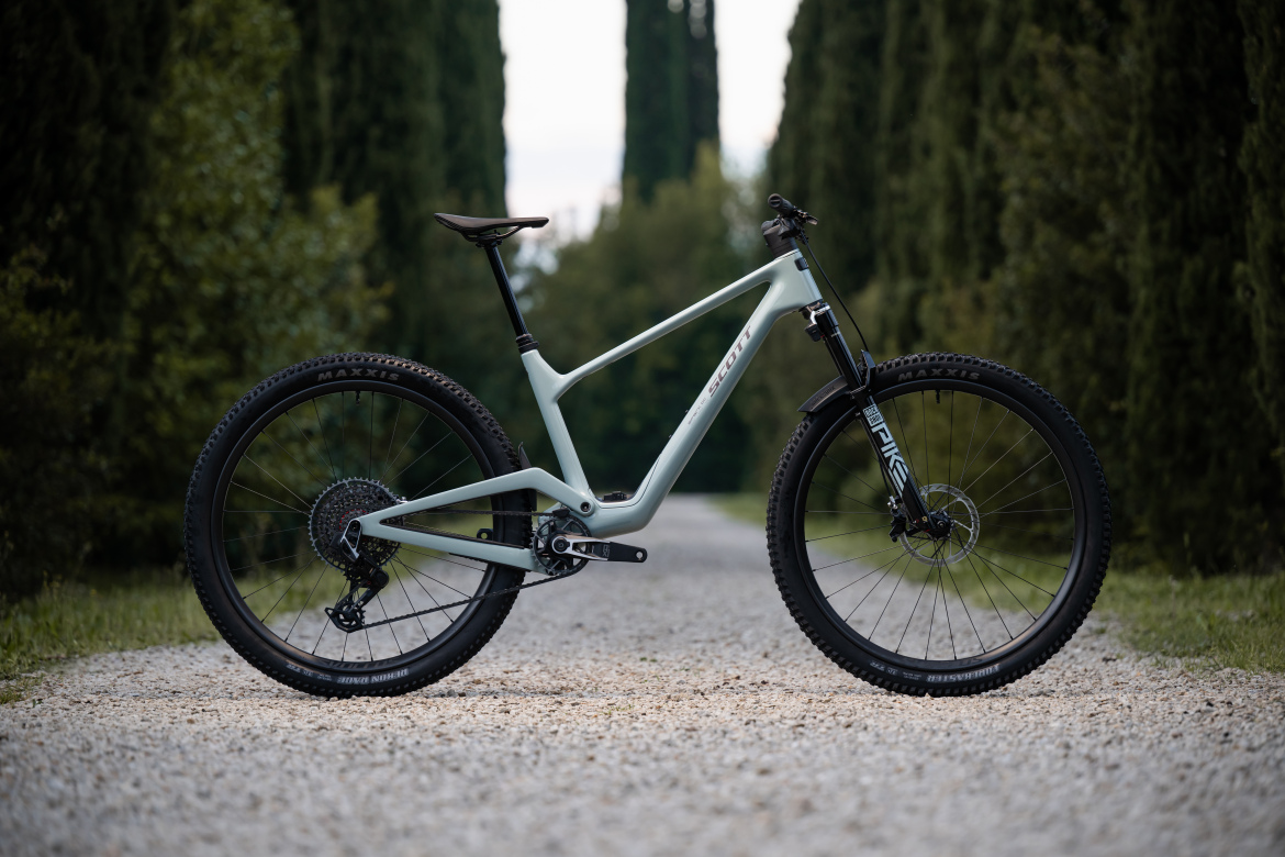 A side view of a modern mountain bike positioned on a gravel path, surrounded by tall cypress trees. The bike features a sleek, light green frame, black wheels with thick tires, and a suspension fork. The background is lush and greenery suggesting an outdoor setting.
