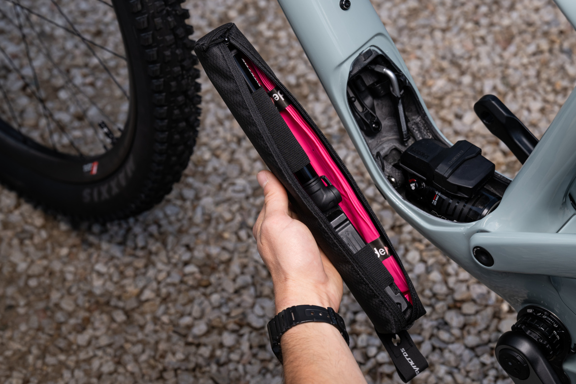 A person holding a black and pink tool case, designed to fit inside a bicycle frame storage compartment. The bicycle is partially visible, with a large tire in the background, and the storage area reveals tools and a battery inside the frame. The ground is covered with small gravel stones.