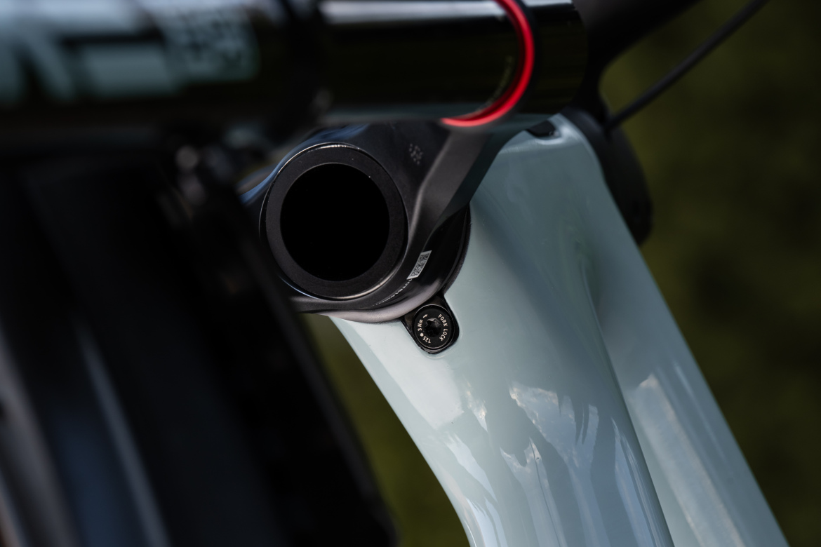 Close-up view of a bicycle frame, highlighting the bottom bracket area with a black casing and a visible screw. The frame has a light blue finish, and there are reflections on its surface, set against a blurred green background.
